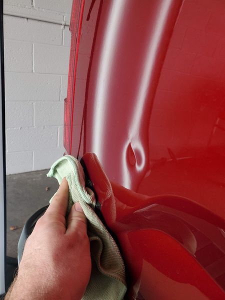 Person's hand wiping a red car's dent with a green cloth. A white wall is in the background.