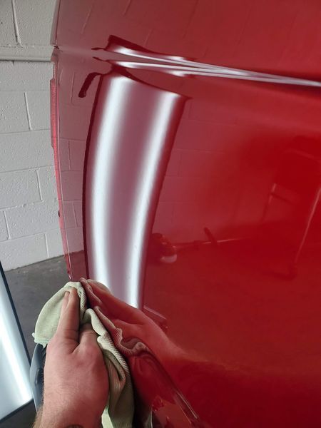 Hand holding a cloth on a red car door, reflecting a dent with white lines.