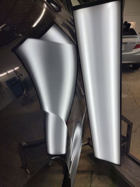 Black car door with a large dent reflecting a long, bright light.