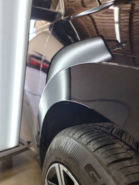 Black car's fender dent being repaired. Reflective surface, tire visible, repair shop setting.