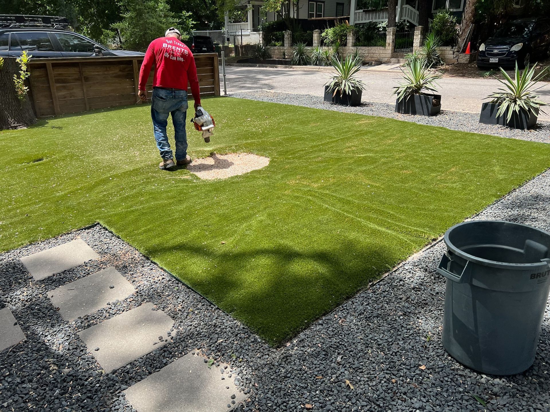 A person in a red shirt uses a power tool on a patch of artificial turf in a landscaped yard with stone pavers and gravel.