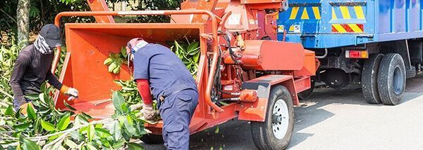 Two workers feeding a wood chipper. The orange machine is next to a blue truck.