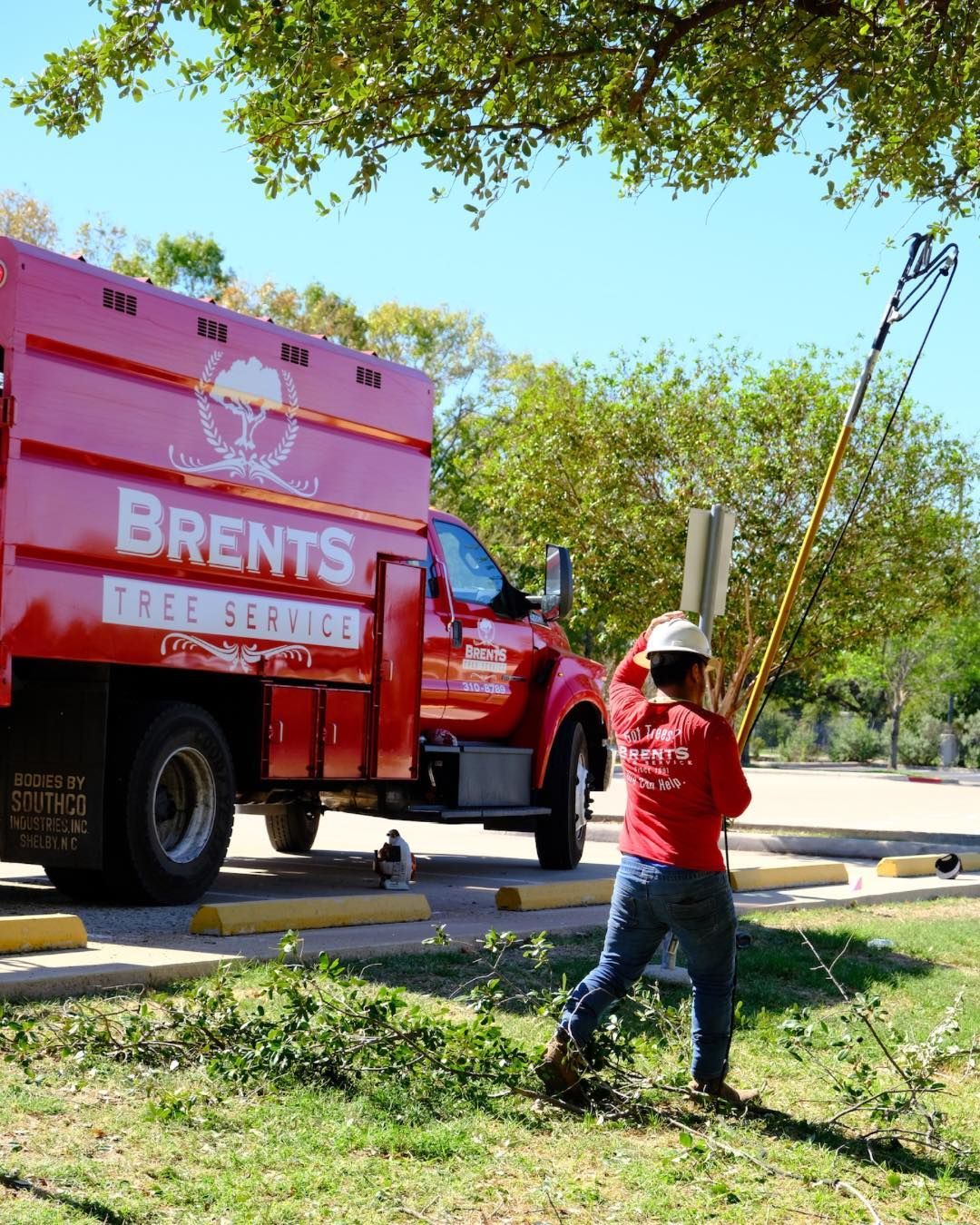 A person in a red uniform uses a pole saw to trim a tree beside a red Brents Tree Service truck parked on a sunny day.
