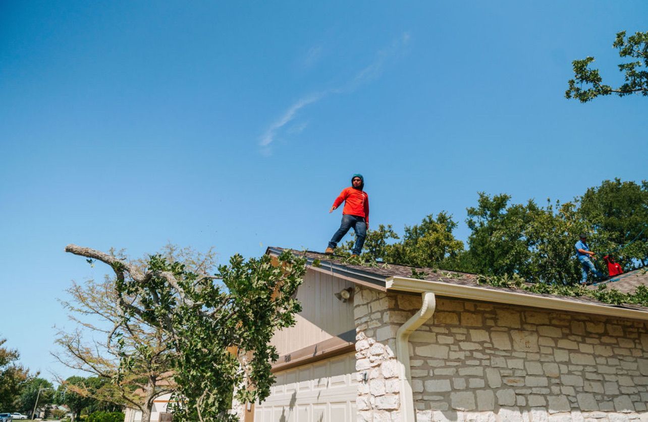 A person in a red shirt walks across a residential roof, with a second person visible near the edge against a blue sky.