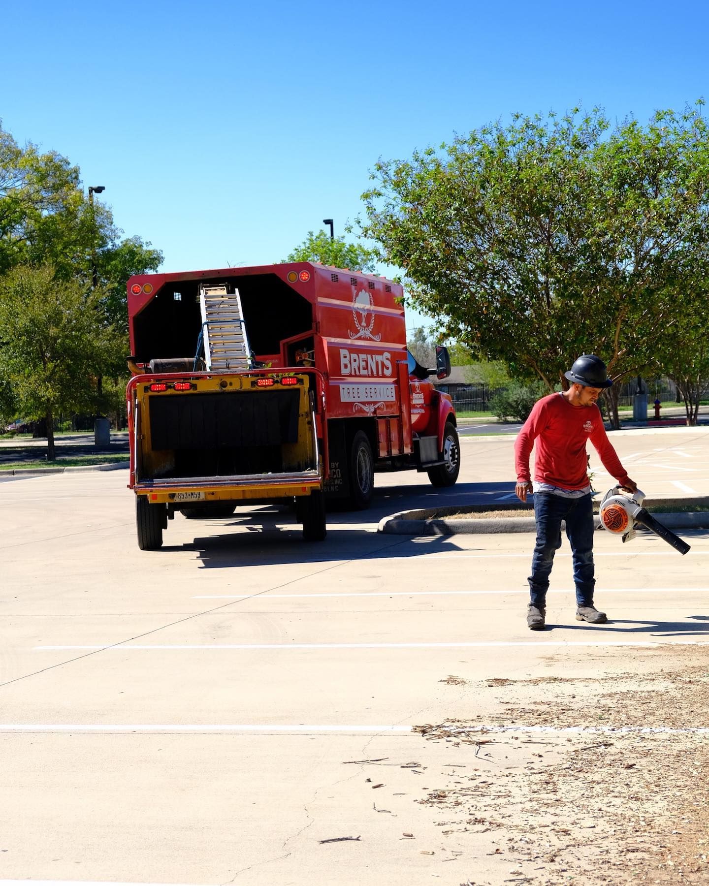 A worker in a red shirt uses a leaf blower in a parking lot next to an orange truck labeled 