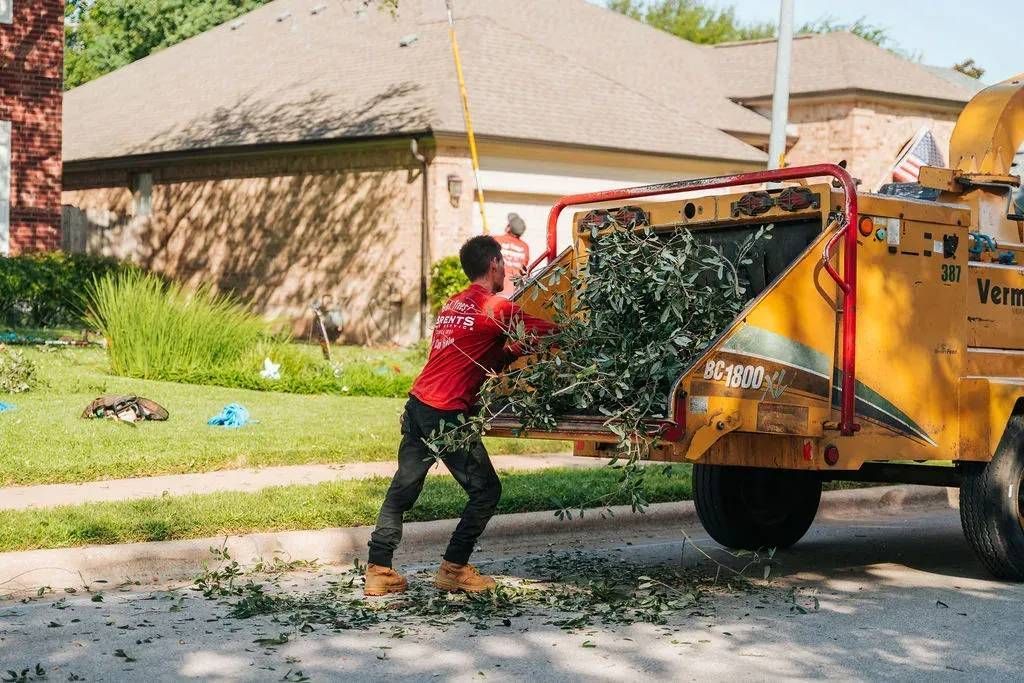 Man in red shirt feeding tree branches into a yellow wood chipper on a sunny suburban street.
