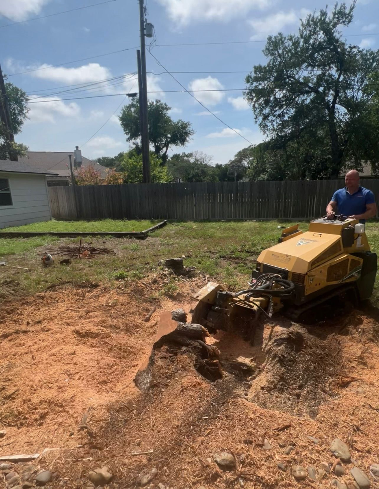 A person operates a yellow stump grinder in a grassy backyard, reducing a tree stump to wood chips in the soil.