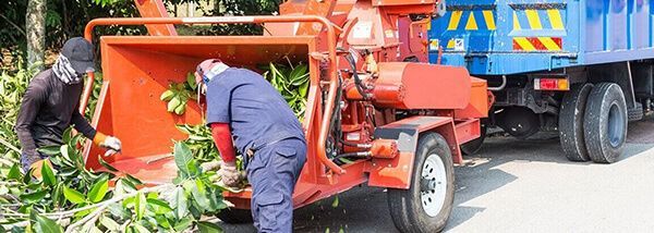 Two workers feeding a wood chipper. The orange machine is next to a blue truck.