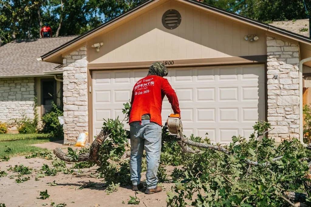 Man in red shirt using a chainsaw to cut tree branches in front of a house with a white garage door.