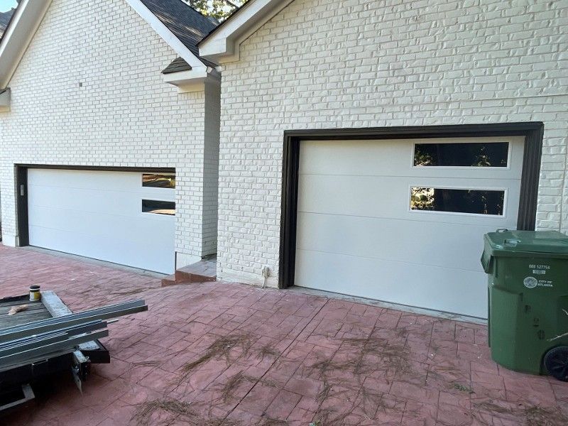 Two white garage doors on a brick driveway, with a green trash bin beside the right door.