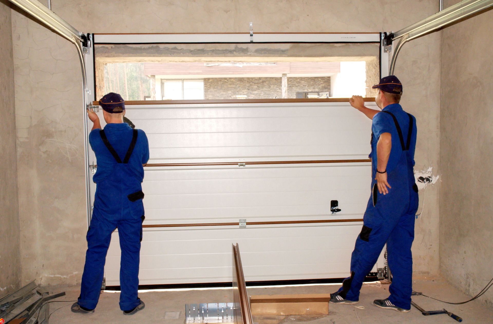 Two workers in blue overalls lift a garage door into place inside a garage opening.