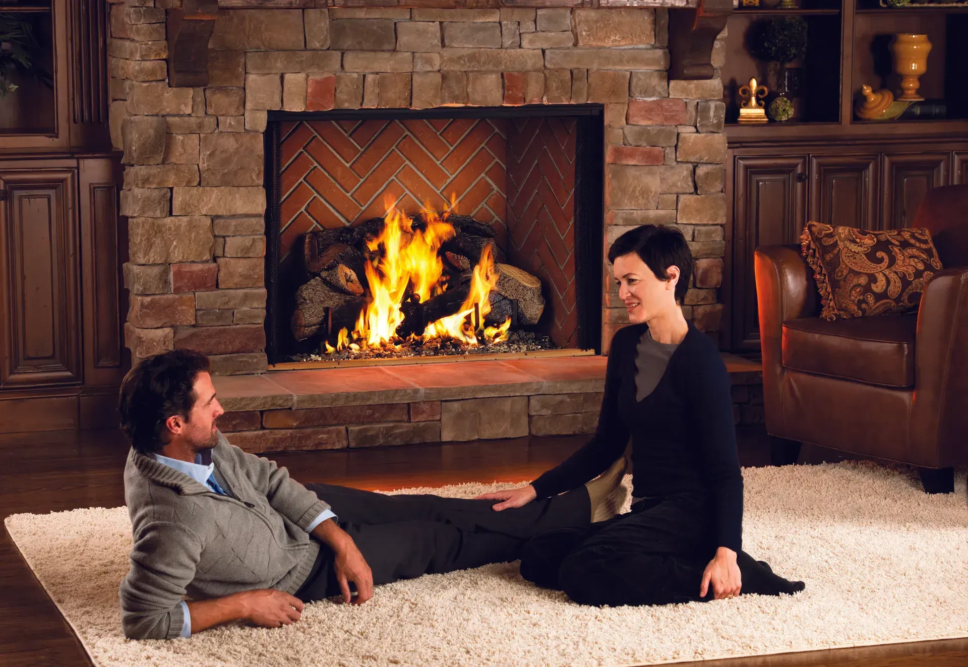 Couple by a fireplace, man lying down, woman sitting, warm fire, neutral rug.