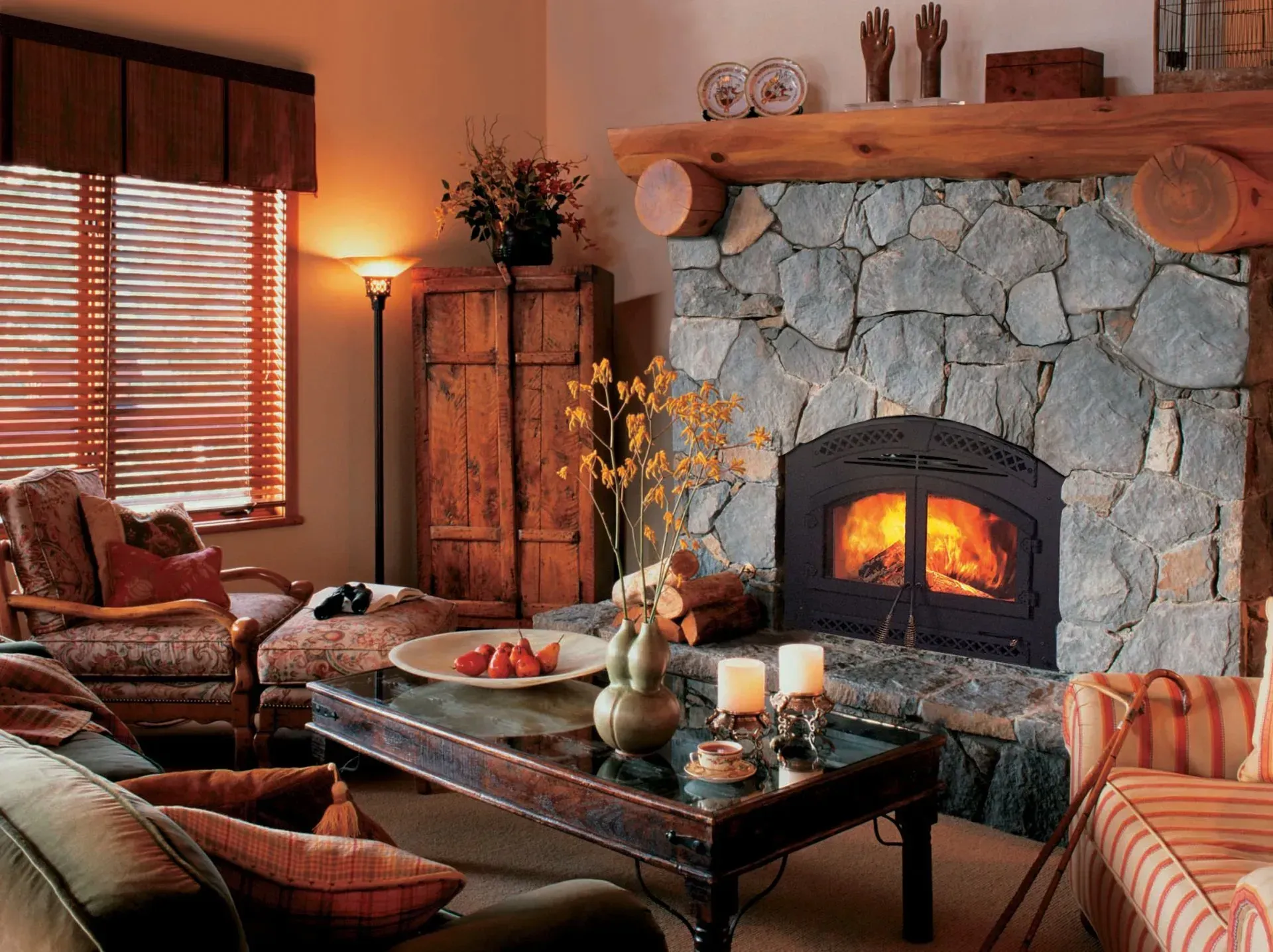 Cozy living room with stone fireplace, lit fire, wood beams, seated furniture, and window with blinds.