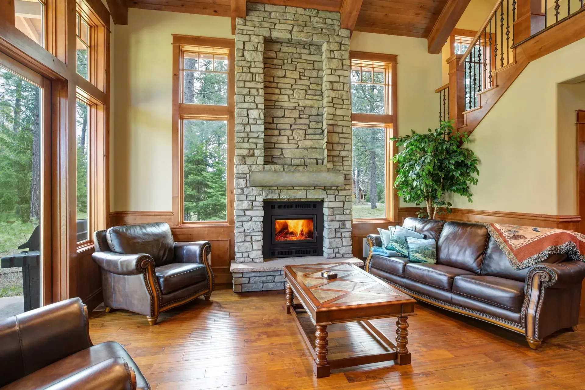 Cozy living room with stone fireplace, leather furniture, and large windows overlooking trees.