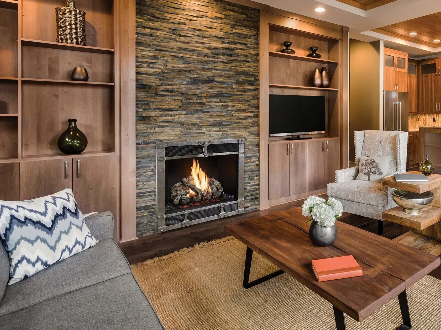 Cozy living room with fireplace, TV, and wooden furniture. Gray sofa, beige rug, and stone accent wall.