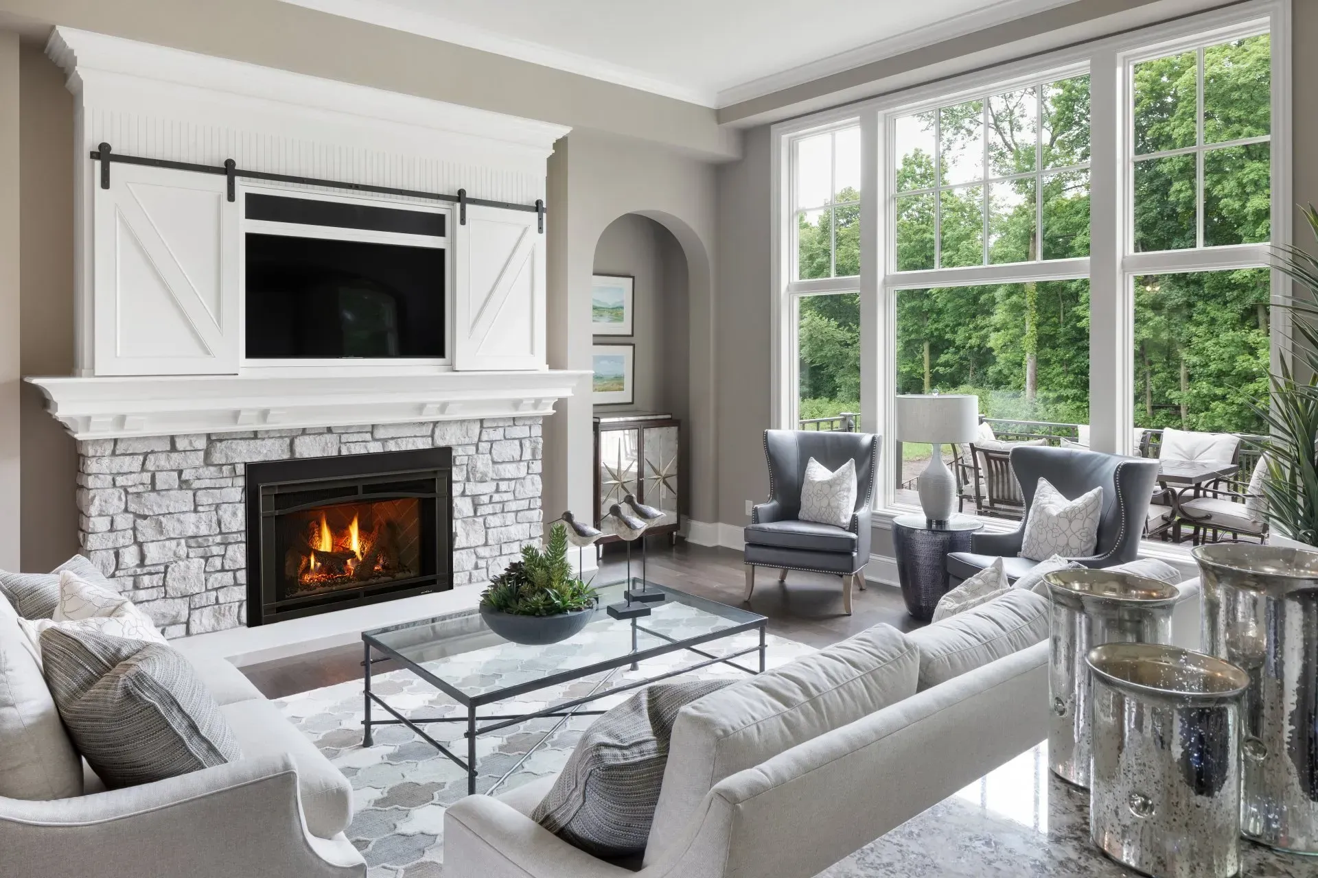Living room with fireplace, light-colored furniture, large windows, and a view of trees.