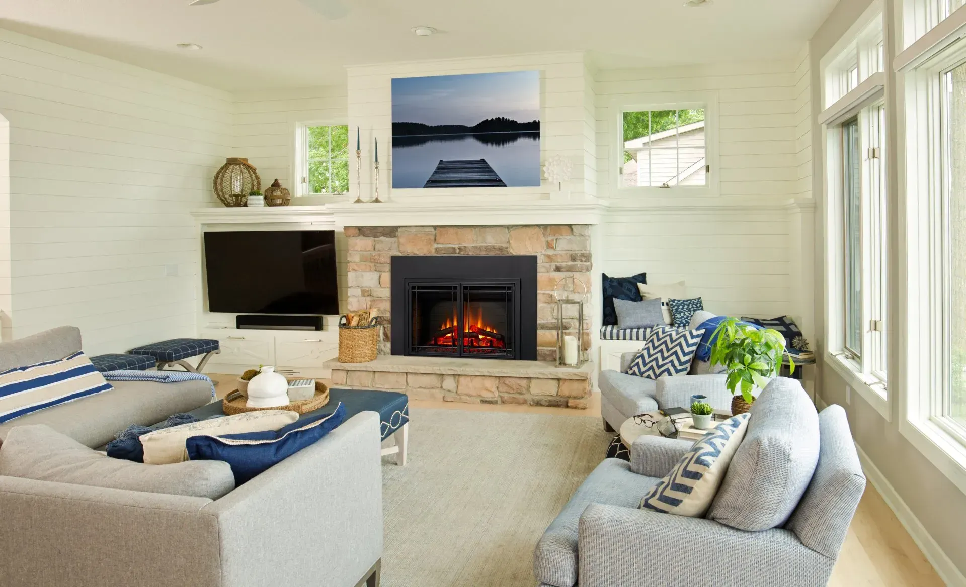 Cozy living room with stone fireplace, gray sofas, and lake art. White walls, large windows, and light carpet create a bright space.