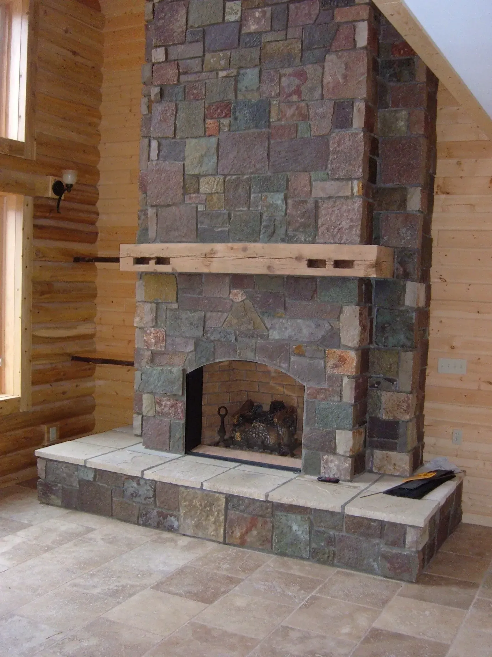 Stone fireplace with wooden mantel and surround in a room with wood-paneled walls and a tiled floor.