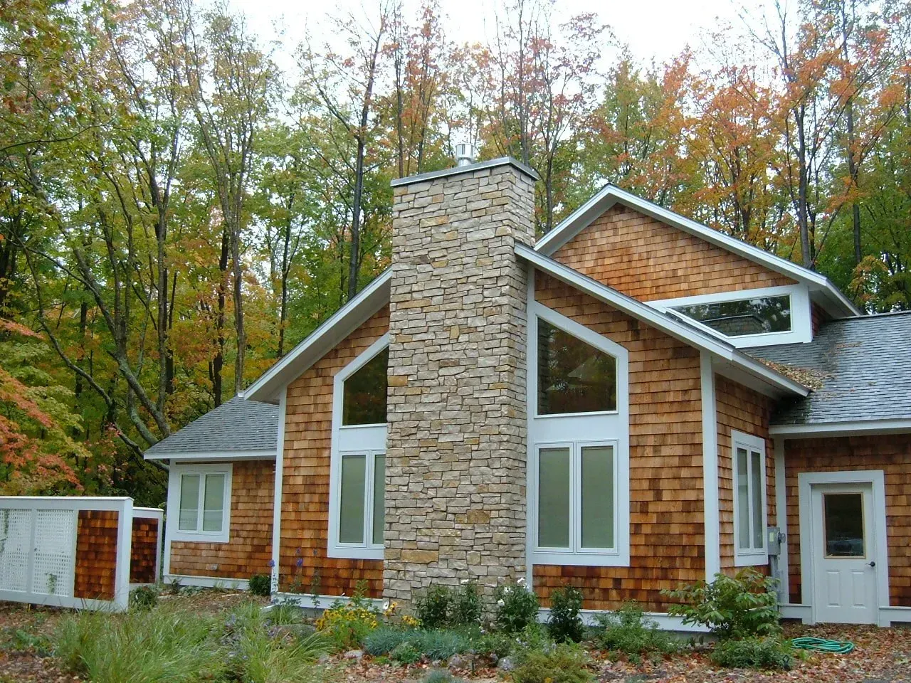Tan shingled house with stone chimney, surrounded by trees with fall foliage.