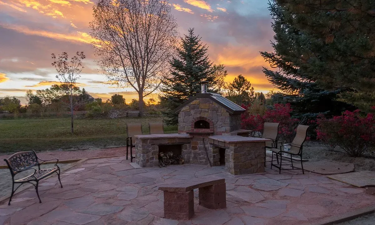 Outdoor stone pizza oven and patio at sunset, with seating and colorful sky.