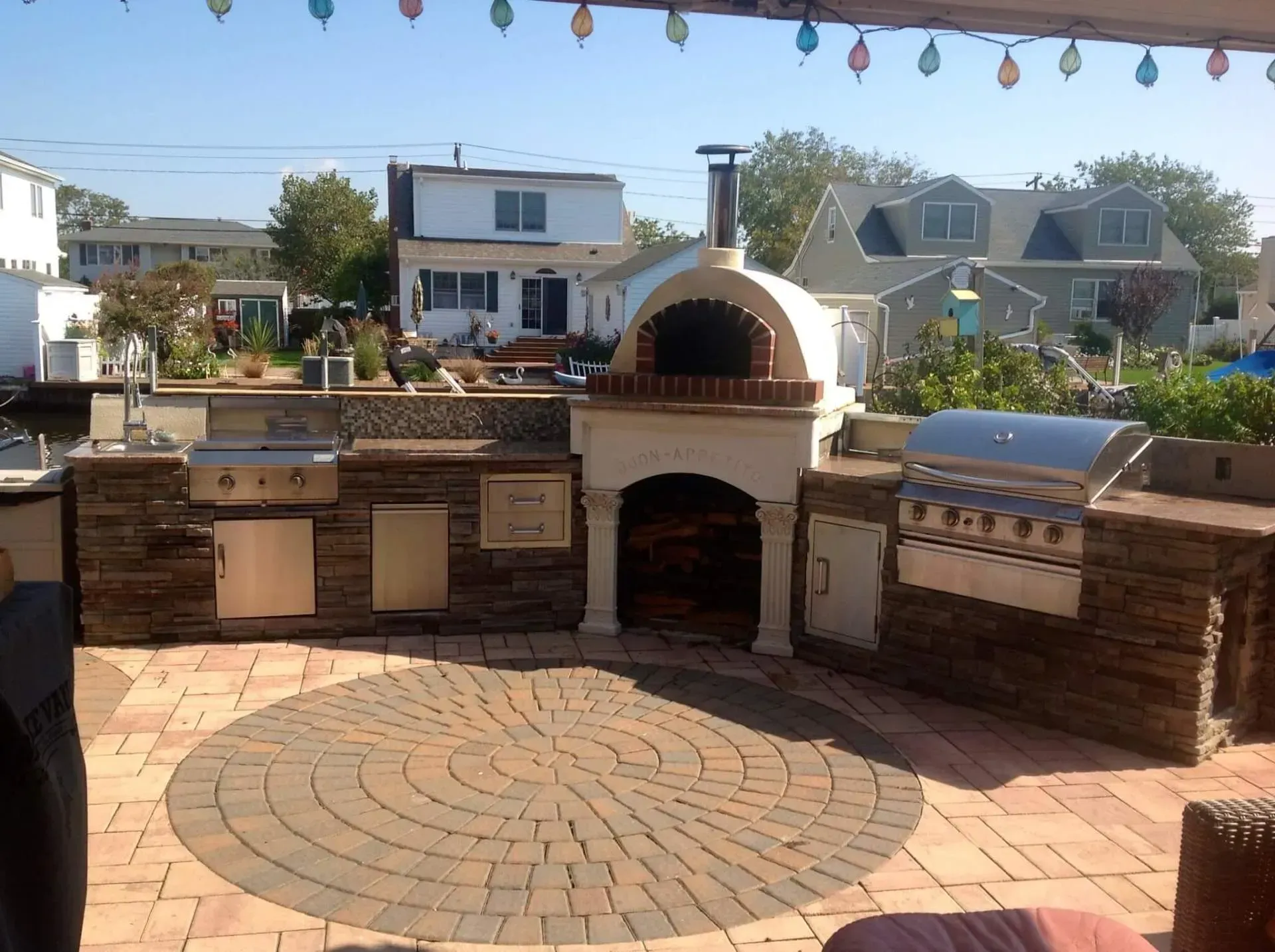 Outdoor kitchen with brick oven, grill, and stone counters on a brick patio near houses.