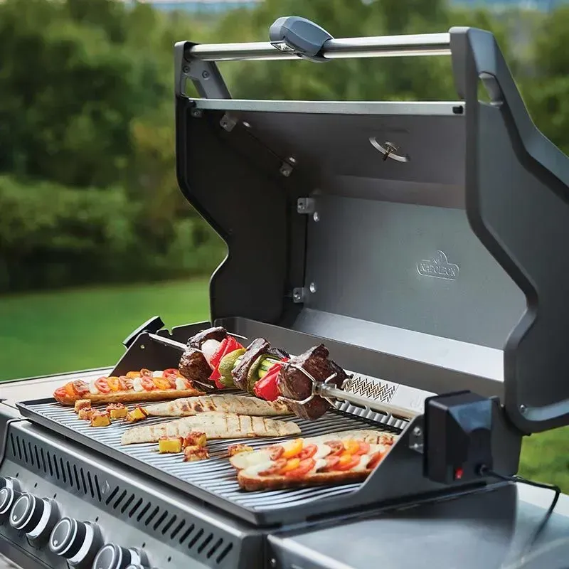 A charcoal grill with open lid, grilling skewers and bread. Outdoors, greenery in the background.
