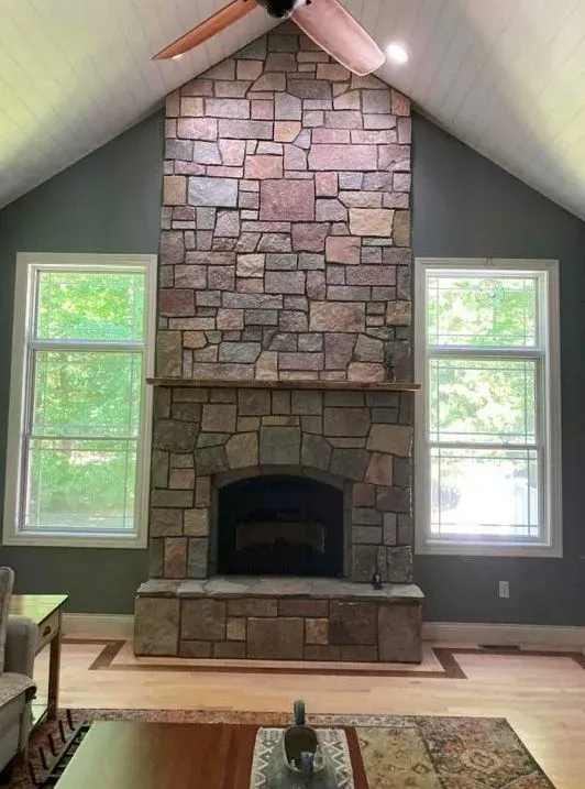 Stone fireplace between two windows in a room with hardwood floors and a high, angled ceiling.