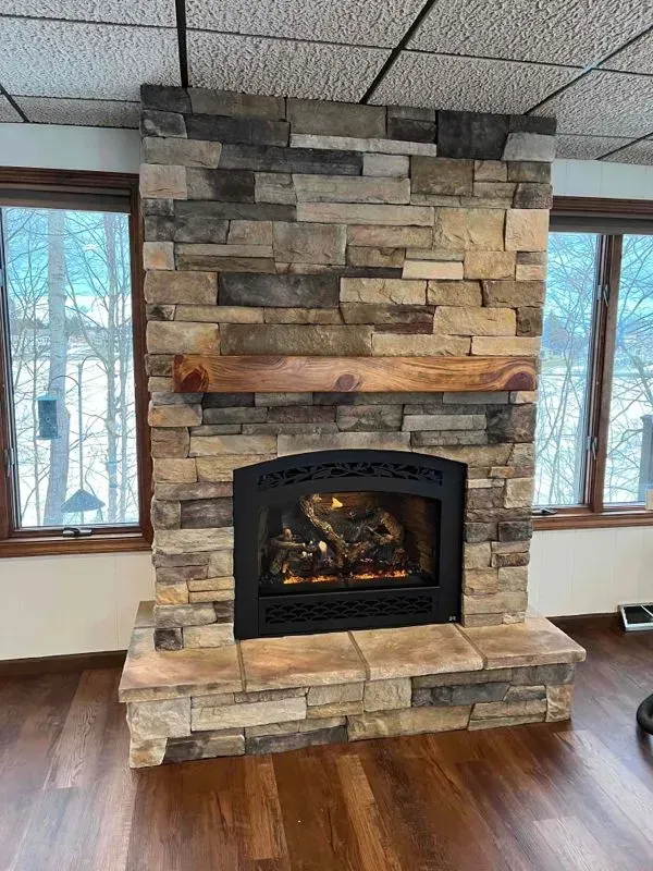 Stone fireplace with a wooden mantel, flanked by windows and dark wood flooring.