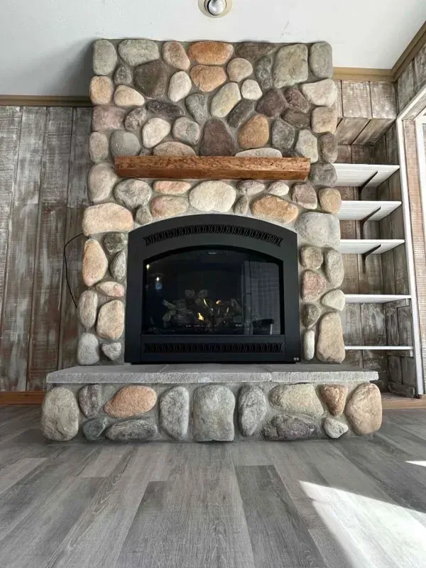 Stone fireplace with black firebox, wooden mantle, built-in shelves, and gray wood flooring.