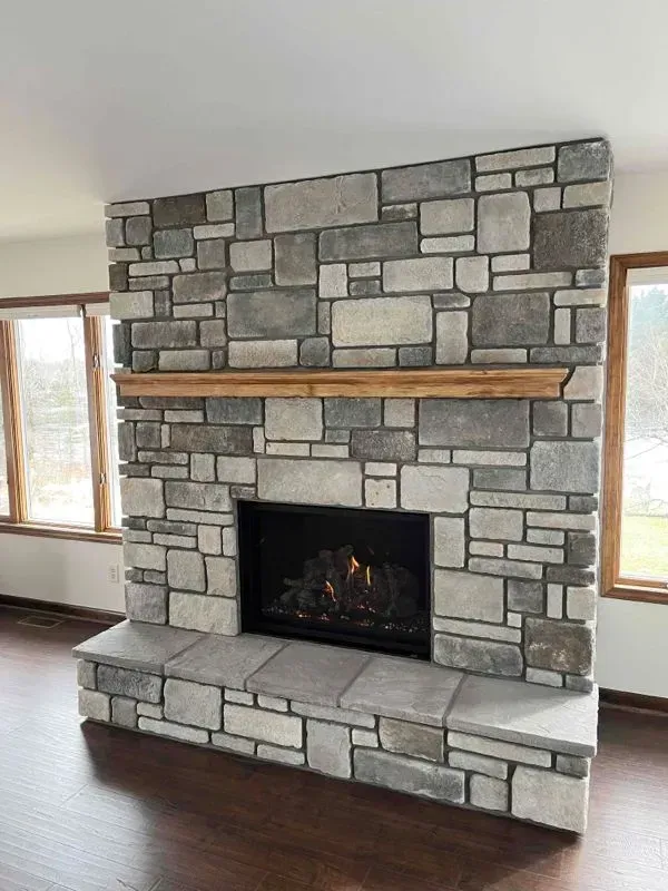 Stone fireplace with gas fire, wooden mantle, and dark flooring in a well-lit room with windows.