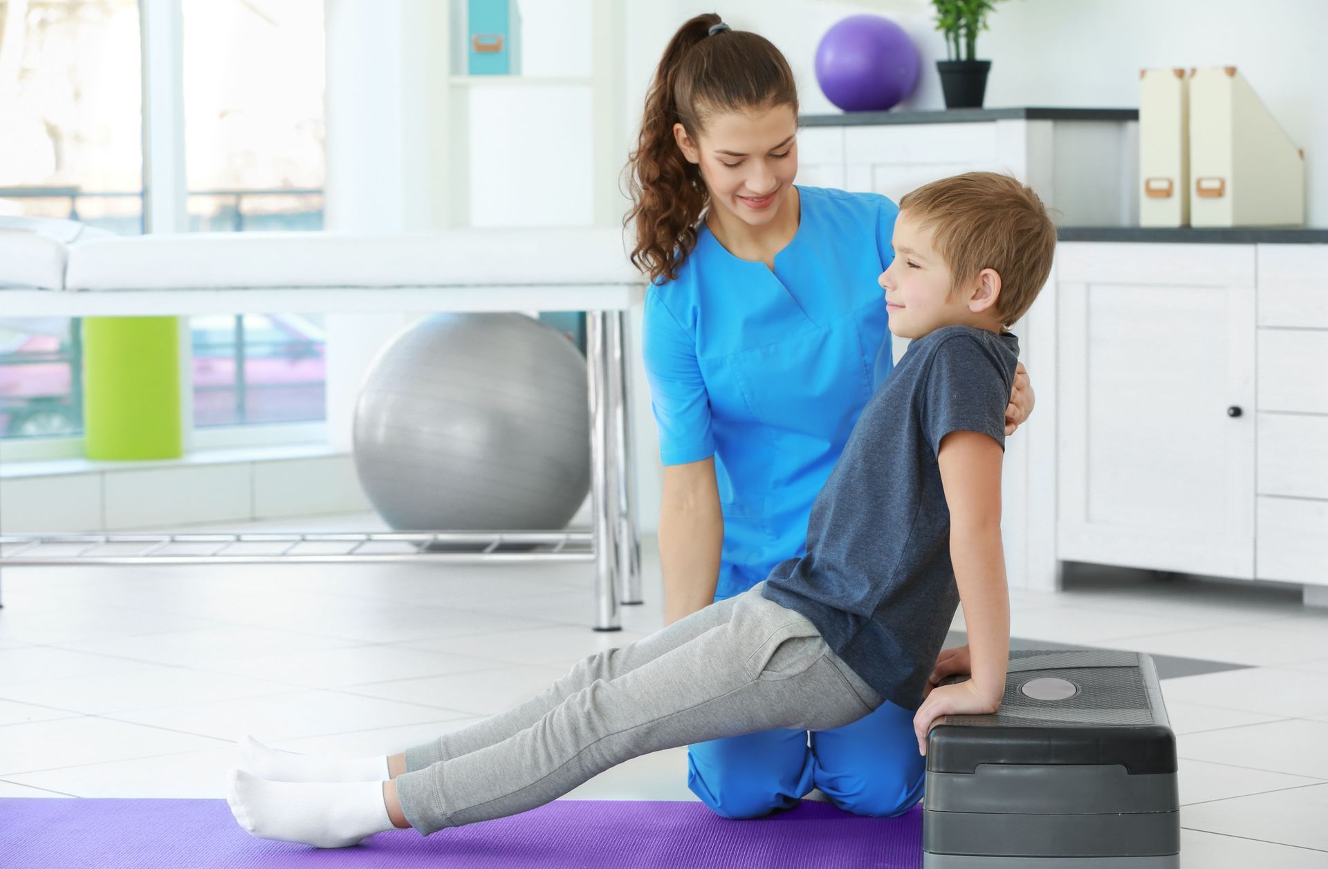 Woman assisting child in a bright gym, with exercise ball and yoga mat nearby.