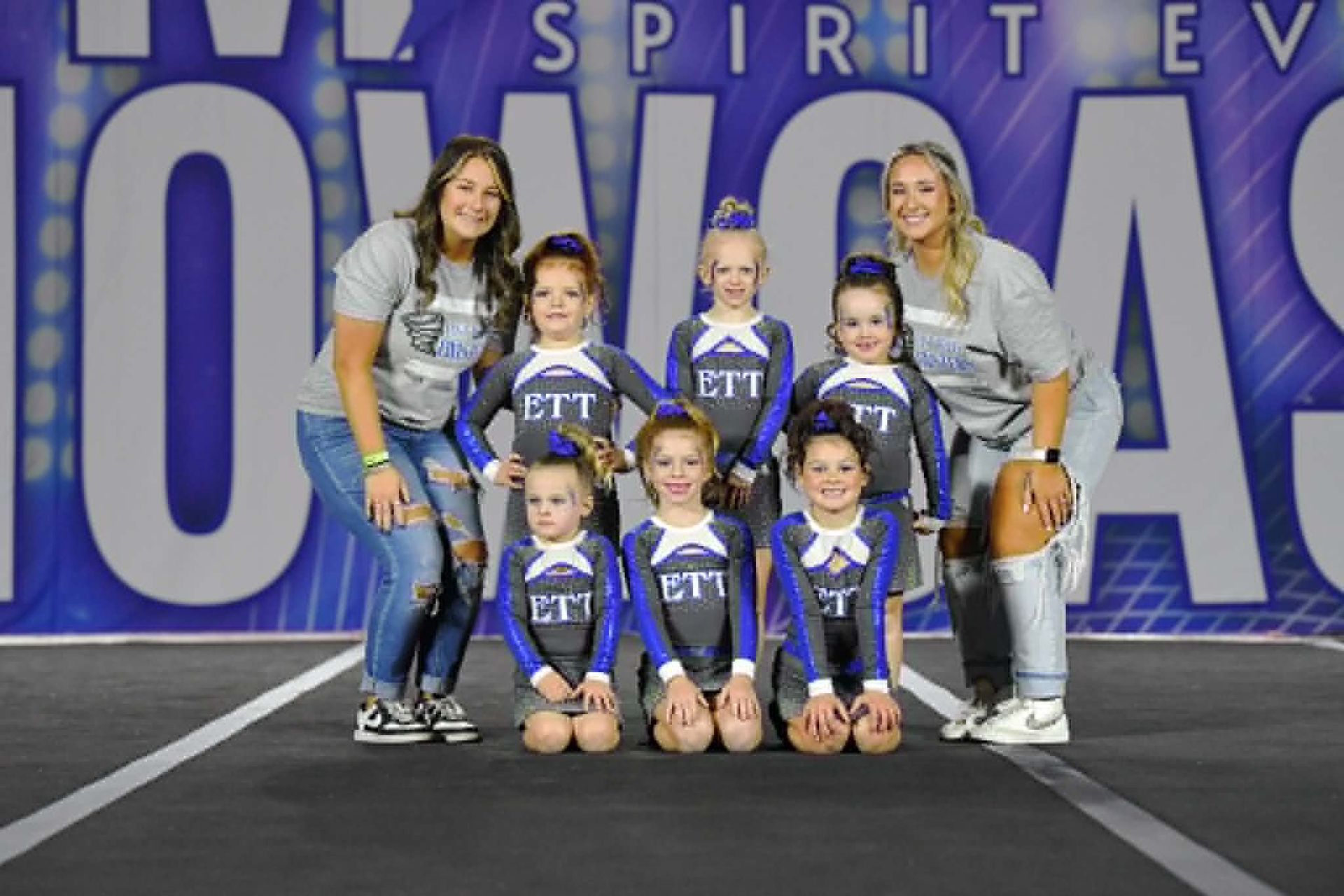 Cheer team posing on a mat in blue uniforms with two coaches in front of a purple event backdrop