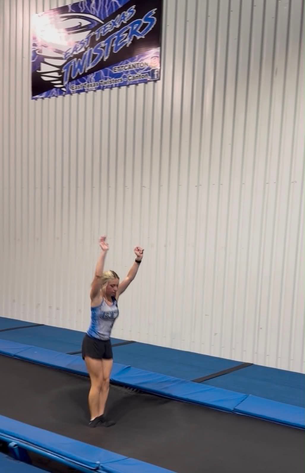 Gymnast standing on a tumbling mat with arms raised in a gymnasium.