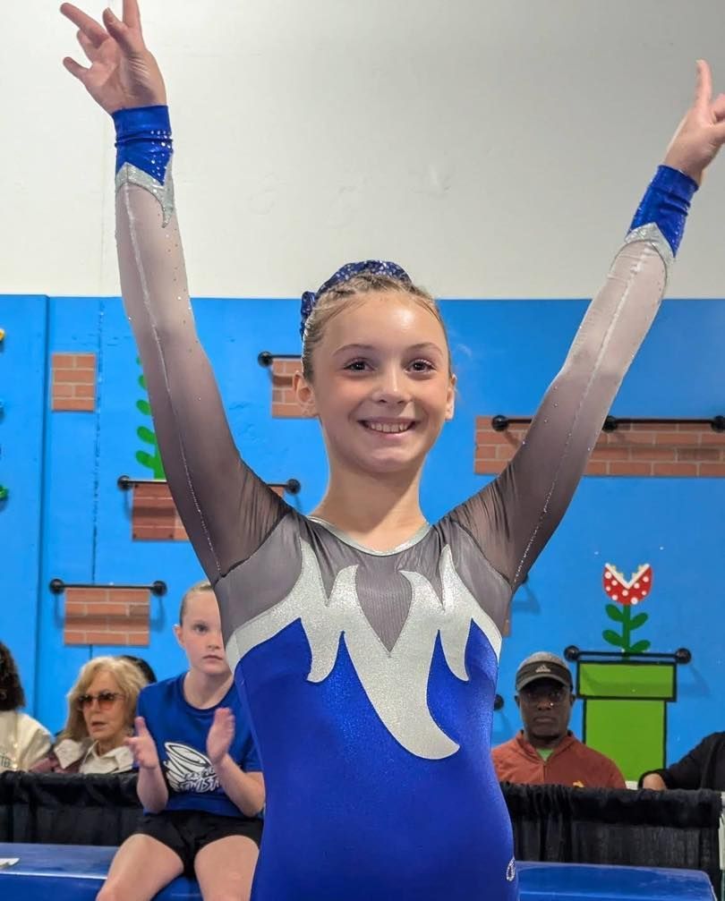 Gymnast in blue leotard smiling with arms raised on a competition mat