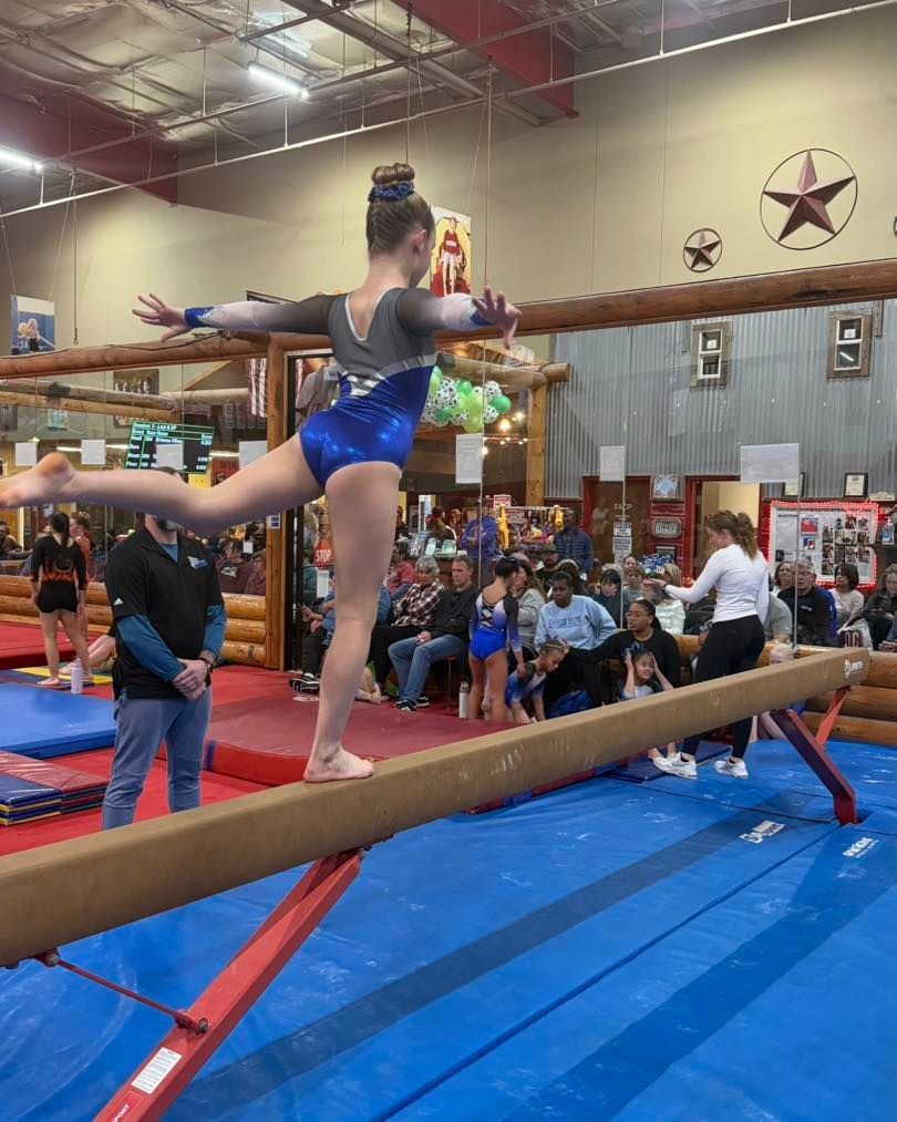 Gymnast balancing on a beam in a blue leotard during a competition inside a gym.