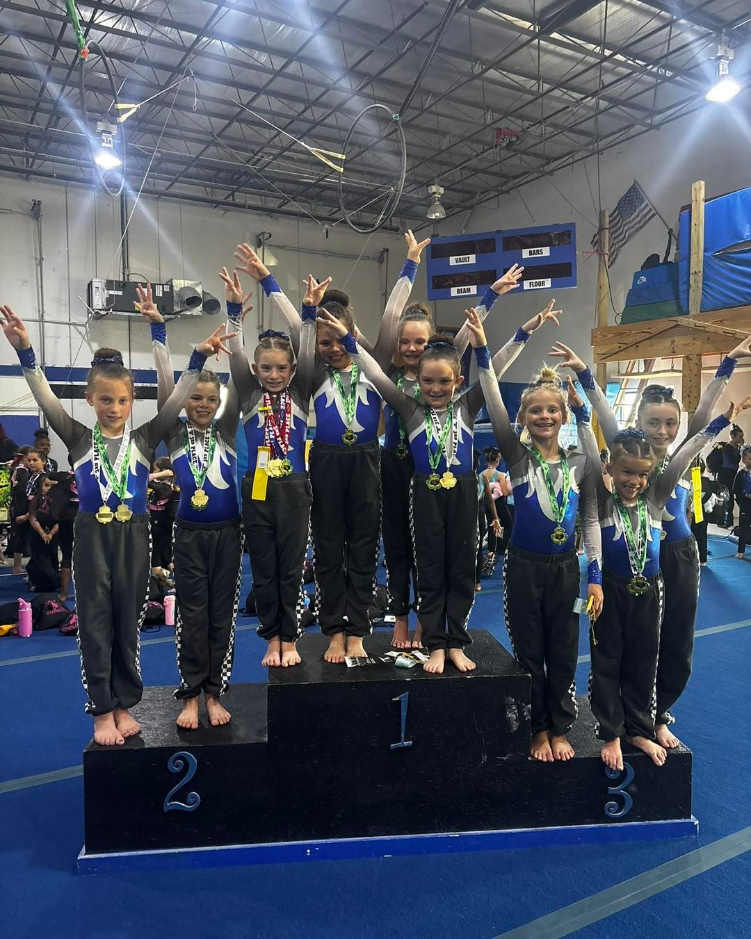 Gymnastics team on a podium, smiling with arms raised, in a gymnasium with medals and trophies behind them.