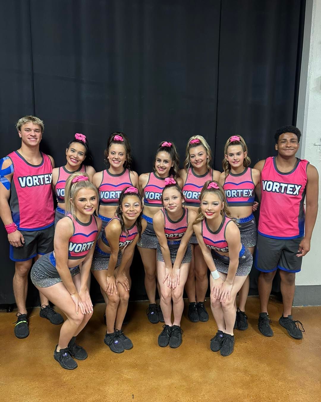 Group of cheerleaders in pink-and-blue “Vortex” uniforms posing indoors on a stage.