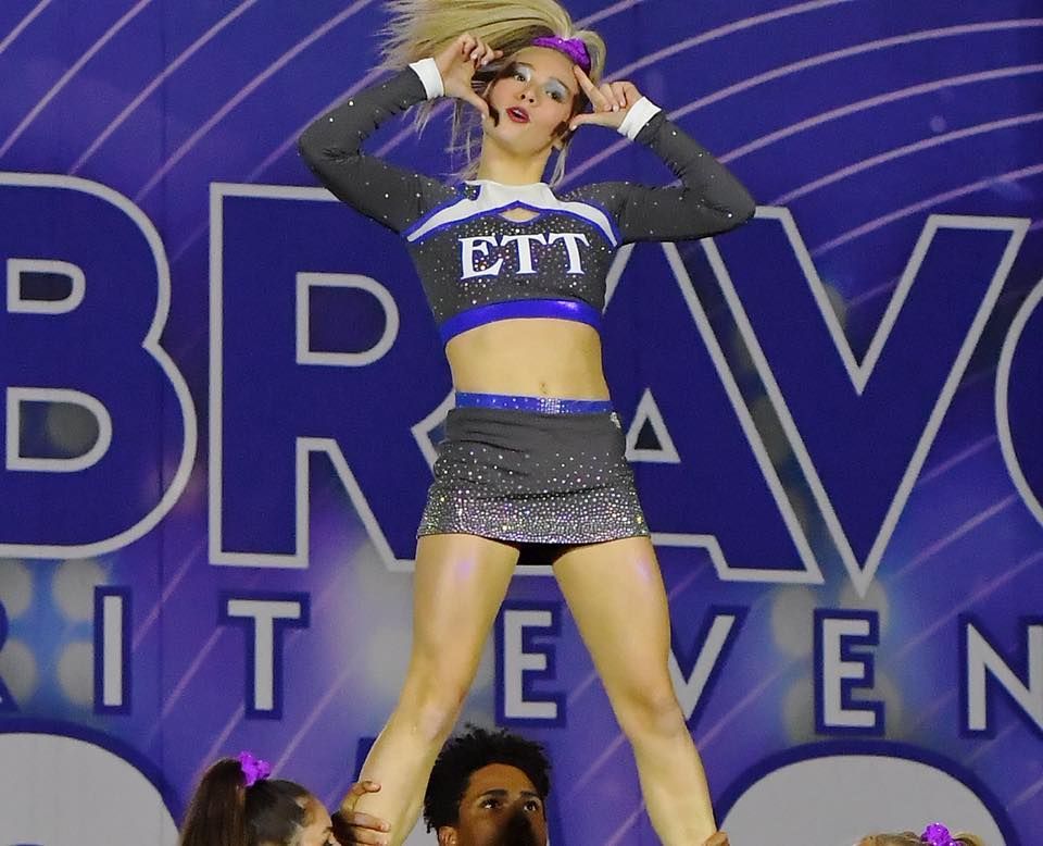 Cheerleader in blue-and-silver uniform posing on stage with arms raised before a BRAVO backdrop