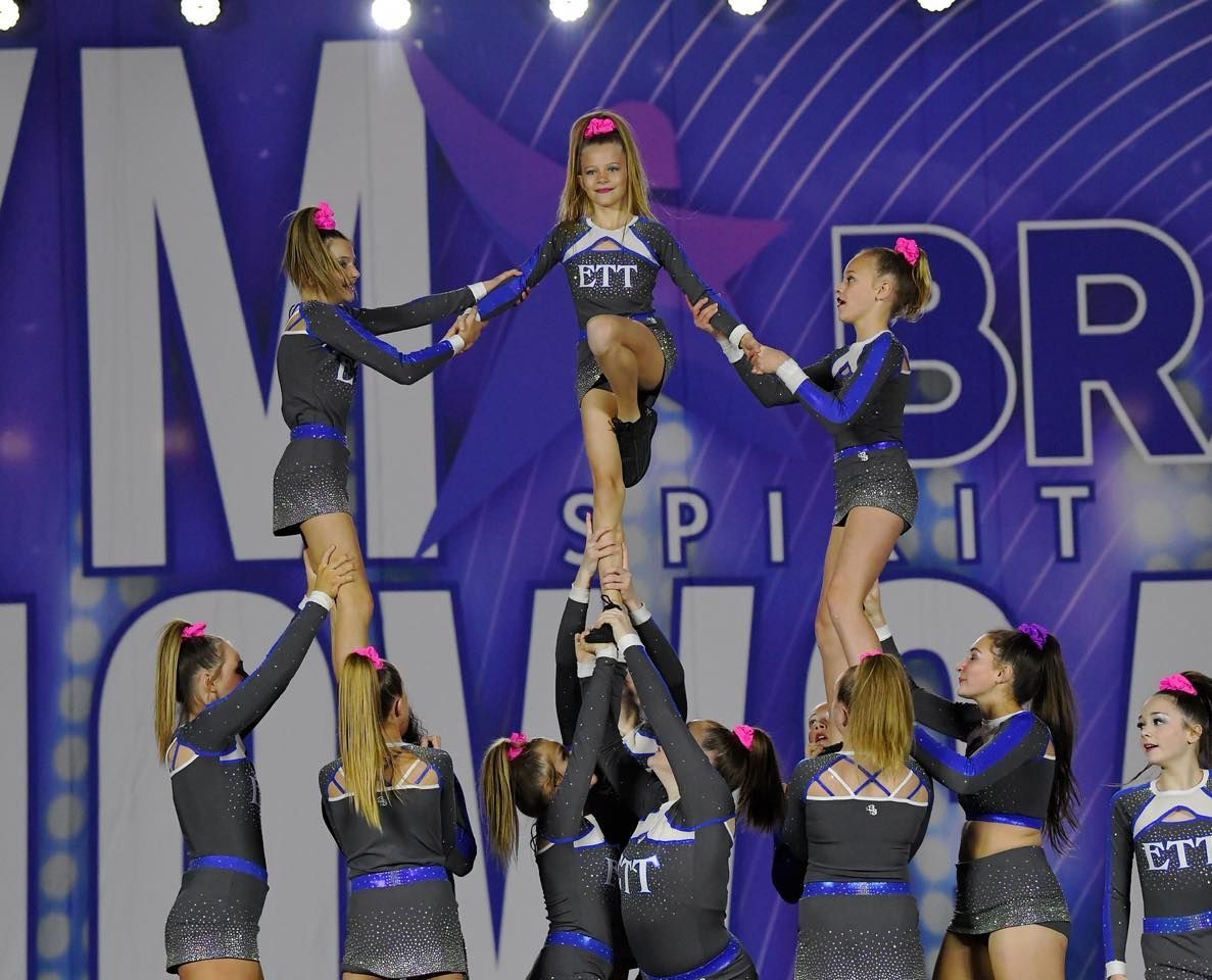 Cheer squad performing a stunt on stage, lifting a girl in a black and blue uniform under purple lights