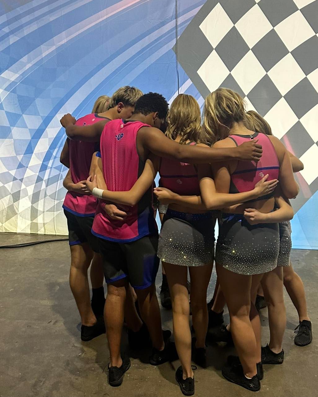 Group of dancers huddled together backstage, arms around each other, wearing pink tops and glittery shorts.