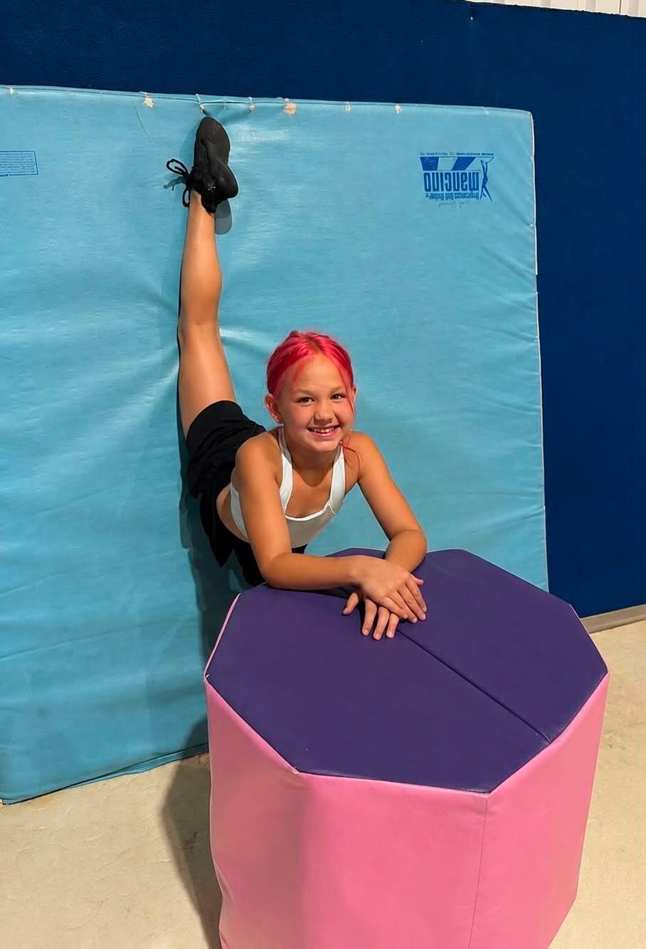 Person posing in a split on a padded gym mat, smiling behind a purple octagonal block.