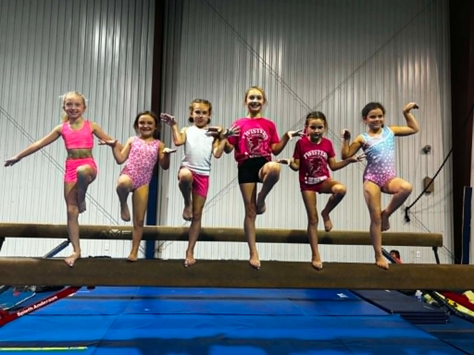 Six children balancing on a beam in a gym, smiling and posing with arms outstretched.
