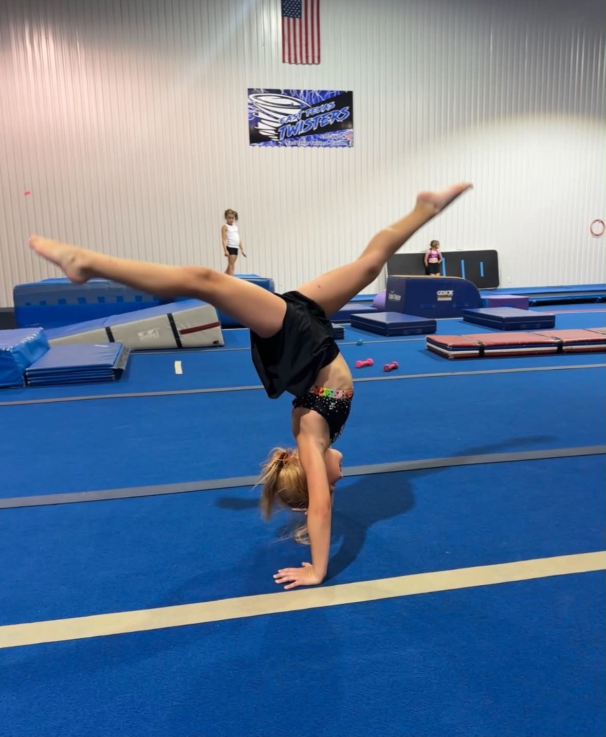 Gymnast doing a one-handed handstand on blue mats in an indoor gym.