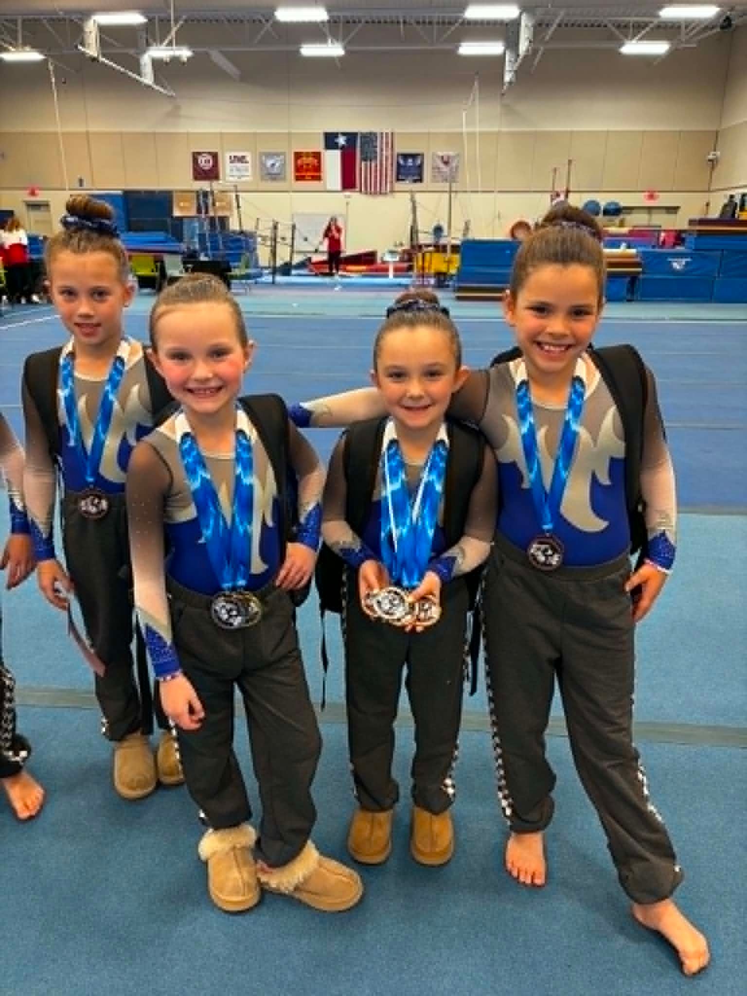 Four gymnasts in blue medals pose on a gym floor after a competition.