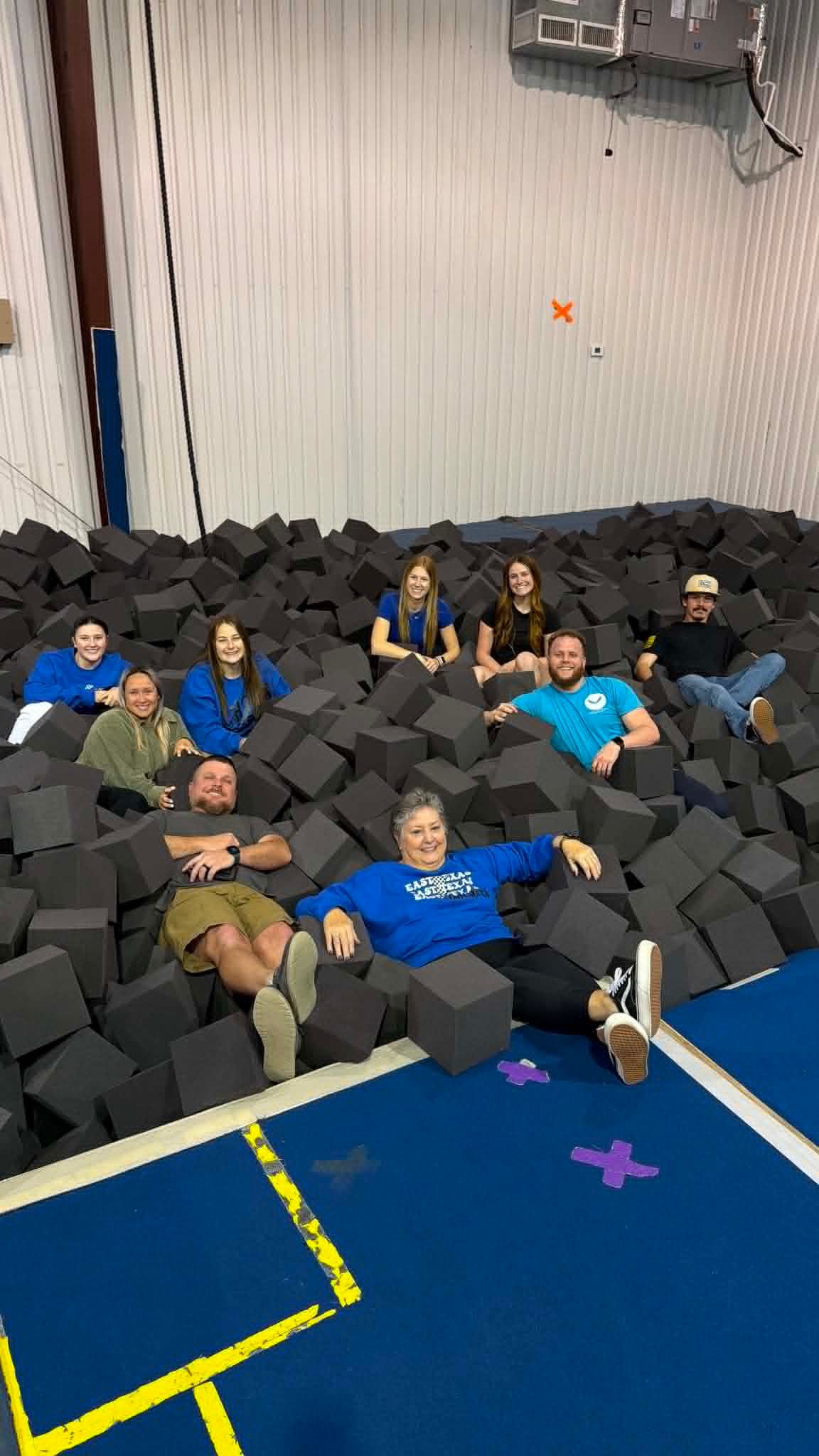 Group lounging on foam pit blocks beside a blue gym mat in an indoor gym