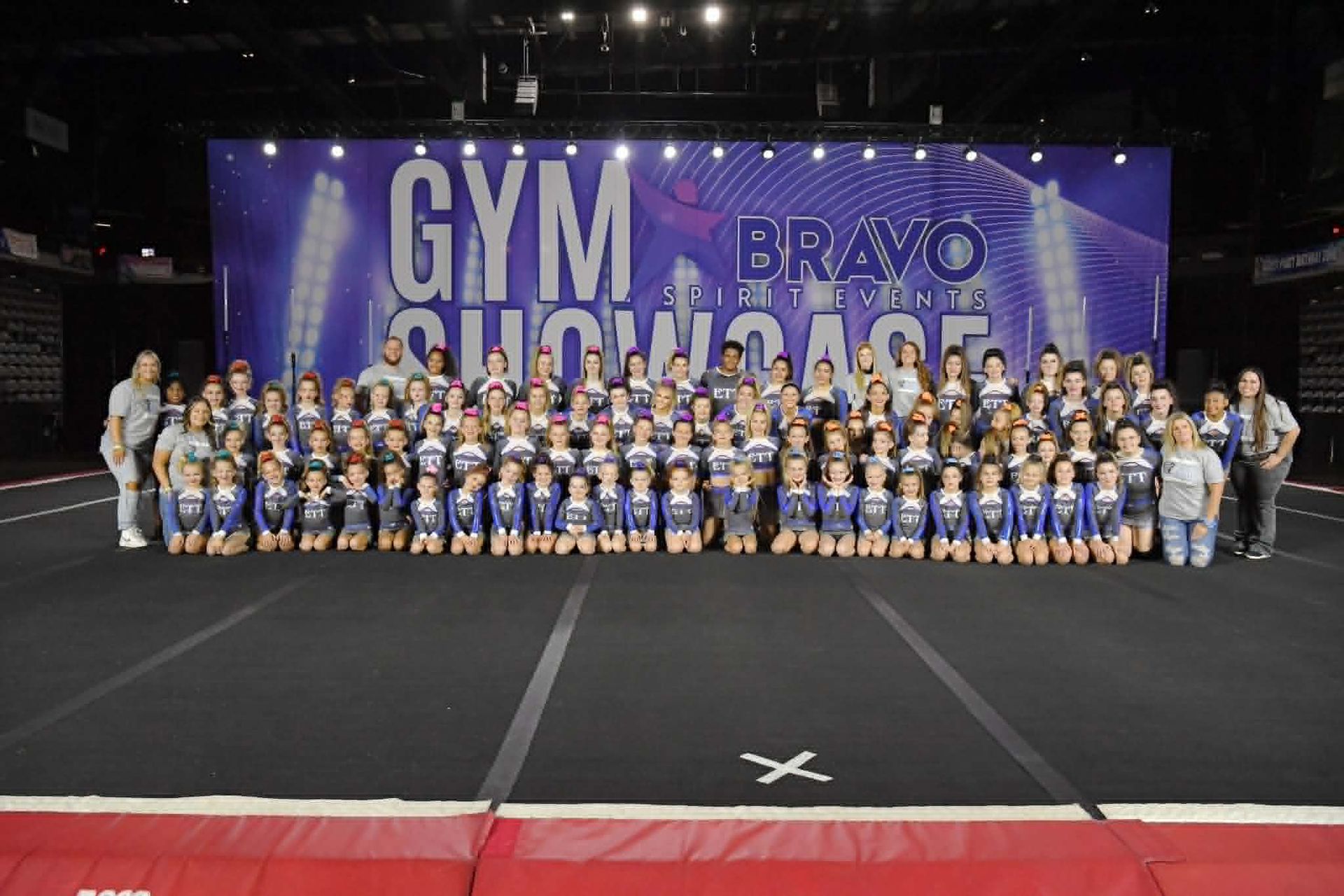 Large gymnastics team posing on a stage in blue-and-white uniforms under a purple 