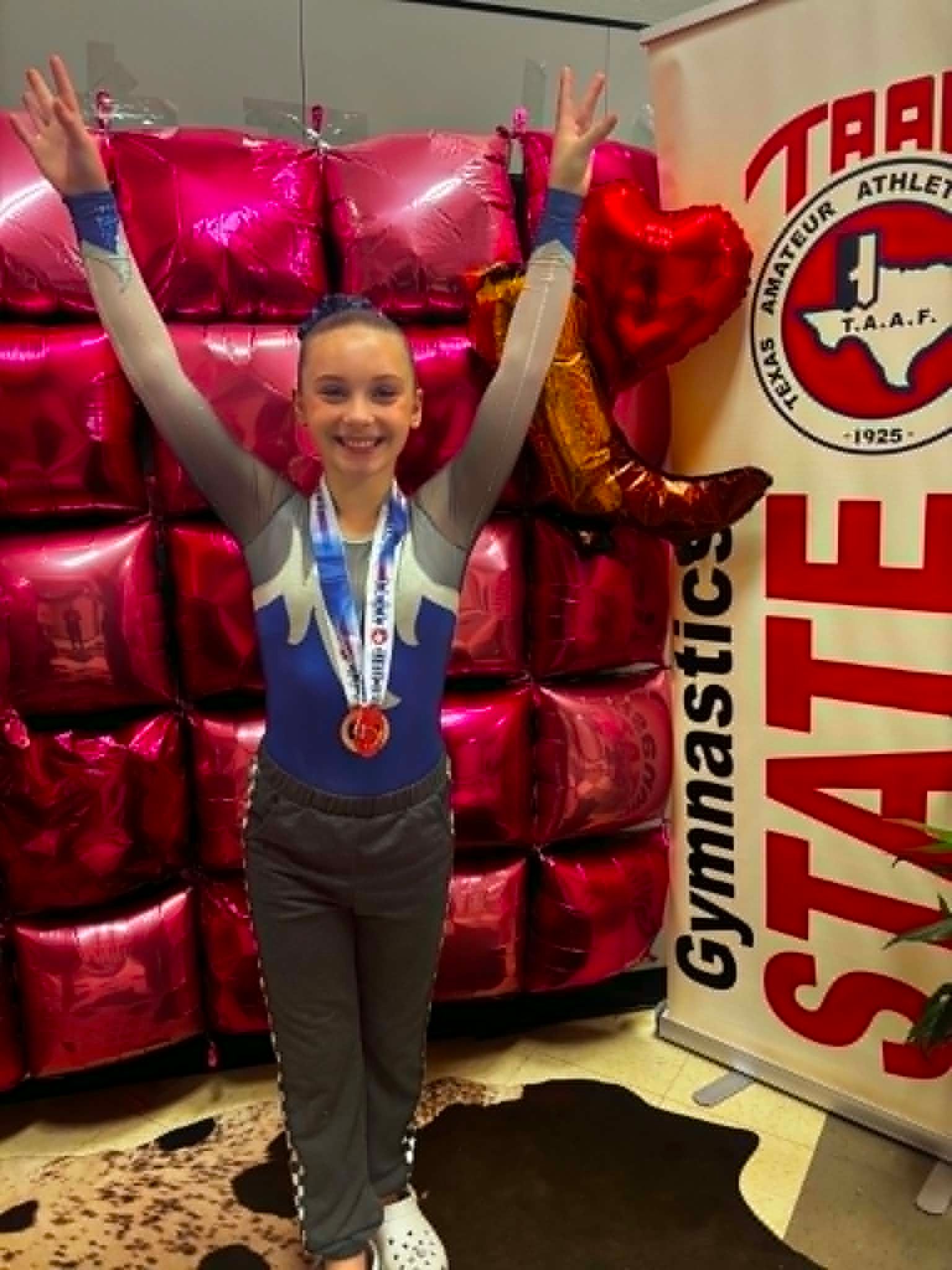 Young gymnast in gray outfit with medal, arms raised, standing by red balloon display and gymnastics banner