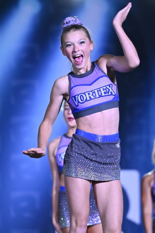 Young cheerleader in a purple “VORTEX” outfit posing on stage with one arm raised.