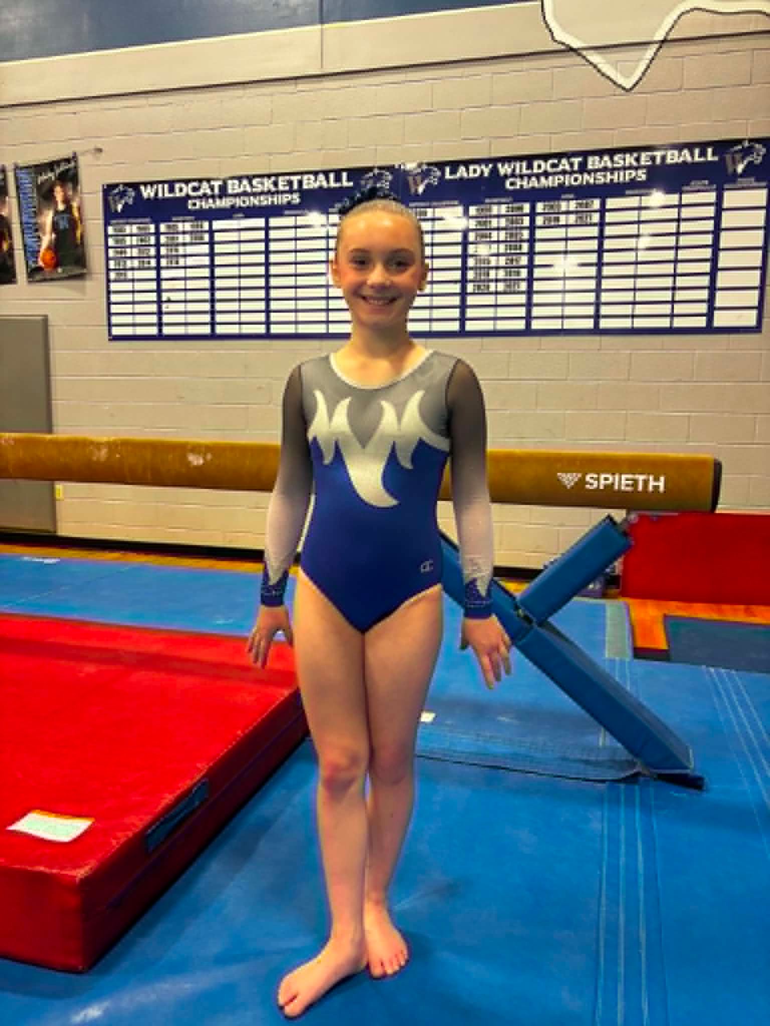 Gymnast in blue leotard standing on mats in a school gymnasium, smiling in front of basketball banners.