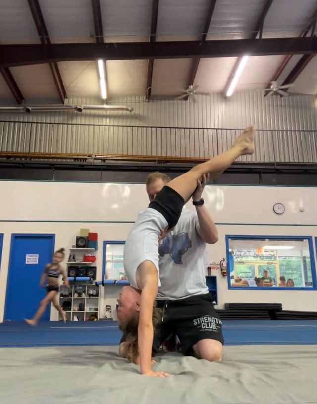 Gymnast balancing upside down on a trainer’s shoulders in a gymnasium